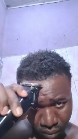 The video shows a close-up of a dark-skinned man using electric clippers to cut his hairline in front of a light purple wall. He looks directly at the camera while holding the black clippers close to his forehead, trimming his short, curly black hair. The background includes part of a tiled wall and ceiling, suggesting the setting is a bathroom or similar space.