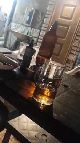 The video shows two men sitting at a table outside. There are drinks and food on the table. One of the men is holding a phone and talking. The other man is out of focus. The video is shaky and the lighting is poor. The area is lit by an overhead bulb.