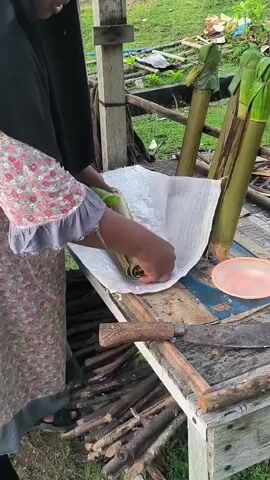 The video shows a person in a black hijab and a floral patterned dress standing at a wooden table. Their hands are busy wrapping a long, green bamboo stalk with a white fabric. To the right of the person, there are other bamboo stalks, some already wrapped in leaves and tied with string. A knife with a wooden handle lies on the table in front of them, along with a small pink bowl. The background shows a rustic outdoor setting with green foliage and more bamboo stalks. The person appears to be in the process of preparing the bamboo for cooking or a similar purpose.