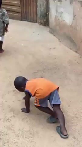 A young child in an orange tank top and gray and orange shorts is on his hands and feet, crawling on sandy ground outdoors near a building. The child briefly shifts to standing, then begins walking away. Two other children are visible in the background, walking away. The child who crawls is wearing slides.