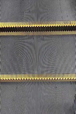 The video shows the steps of a moving escalator. The steps are metal with yellow safety lines. The camera is looking down at the escalator. The escalator is moving upwards. The environment appears to be a public place such as a mall or airport. There are no people visible in the video.