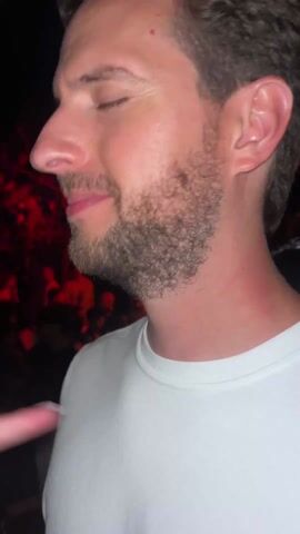 A man and a woman are at a crowded outdoor event at night. The man is white, with brown curly hair and a short beard, wearing a white T-shirt. The woman is white, with light brown hair, and she is wearing a necklace. The background shows many people, and lights that alternate between red and blue. The music is loud.