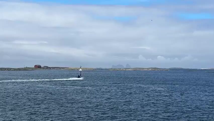 A jet ski is shown being ridden across a body of water. The jet ski is blue and black and is leaving a white wake behind it. The rider is wearing dark clothing and a helmet. In the background, there are several small islands with rocky shores and sparse vegetation. There are also some buildings visible on one of the islands. In the far distance, there are mountains that are partially obscured by clouds. The sky is overcast with some patches of blue. The water is a deep blue color.