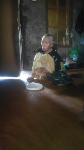 An elderly person with grey hair is squatting on the floor, holding a white paper plate in their right hand. They are wearing a yellow and white patterned skirt and a dark-colored shirt. The person appears to be eating or preparing to eat from the plate. The background shows a room with a rough-textured wall, shelves with items on them, and black plastic bags. A green helmet is visible on the shelves. The lighting suggests an indoor environment, possibly a humble dwelling.