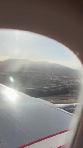 The video shows the view from an airplane window during the daytime.  The sun is high in the sky, creating a glare on the airplane wing, which is visible in the foreground. The wing is white with a red stripe and arrow. Beyond the wing, a landscape stretches out, including a town with white buildings, roads, and brown, semi-arid terrain.  Mountains are visible in the distance, partially obscured by haze or thin clouds. The overall lighting is bright, but the haze softens the distant details.
