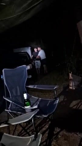 The video shows an outdoor scene at night, possibly a campsite, with a tarp overhead. In the foreground, a dark folding camping chair is visible with a green bottle on it. Two men are standing near the open trunk of a car in the background. One man, wearing a light-colored t-shirt and dark shorts, is leaning into the trunk. The other man, wearing a dark t-shirt and light-colored shorts, stands beside him. The lighting is dim, suggesting it is nighttime.