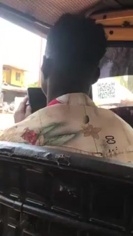 From a low-angle perspective inside a moving vehicle, we see the back of a person speaking on a cell phone.  The person, dark-skinned with short dark hair, wears a light colored shirt with floral print. Outside the vehicle, buildings, people and motorbikes are visible in the background, indicating the vehicle is on a street. The vehicle's interior is dark, with a black, worn-looking seat back in the foreground.  A QR code sticker is visible on a rear window.