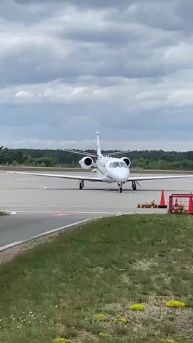 A white private jet, possibly a Cessna Citation, is shown on an airport tarmac under an overcast sky. The aircraft is facing forward, with its engines running, producing an audible hum. It appears to be either stationary or slowly taxiing.

The scene includes a paved runway or taxiway, with green grass and some yellow wildflowers bordering the pavement on the right and foreground. In the background, a dense treeline is visible under the cloudy sky. To the right of the jet, there are orange safety cones and some yellow ground equipment, including a yellow sign with "FOD" partially visible on it, likely indicating a Foreign Object Debris container.

The video was captured during the daytime at what appears to be Skulte Airport in Latvia.