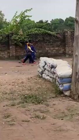 The video shows a few children running and playing in a dirt area near a brick wall and stacks of bagged material, likely cement. The surroundings include trees and a building with a corrugated metal roof.  Later, a goat appears in the distance. The camera seems to be positioned from a slightly elevated point under a canopy or awning, as indicated by the chair with writing on the back.