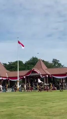 A wide shot captures a scene of cultural significance, likely a celebration or performance, set against the backdrop of a traditional Indonesian building. The building, adorned with red and white banners, boasts a distinctive terracotta roof with a central gable. A large Indonesian flag waves prominently on a tall flagpole in front of the building. In the foreground, on a well-manicured lawn, a group of people, presumably dancers, are engaged in a synchronized performance. They are dressed in colorful and elaborate costumes, suggesting a traditional dance or ritual. As the video progresses, colored flags are introduced, adding to the vibrancy and festivity of the scene. The sky above is a pale blue, dotted with fluffy white clouds, contributing to the overall pleasant atmosphere of the event. A small airplane can be seen in the distance. The camera remains static throughout the shot, focusing on the performance and the surrounding environment.