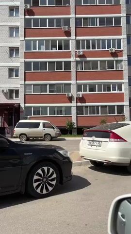 The video begins with a white sedan parked on a paved surface in front of a brick and white apartment building. Air conditioning units are mounted on the building's exterior. The camera then pans to the right, showing the car from the side. The next shot is taken from inside a vehicle, looking out a tinted window at a white sedan. The building is visible in the background, with its windows reflecting the sky. The camera then moves to the rear of the white sedan. The final shot shows two white cars on a road in front of apartment buildings. The sky is clear and blue.