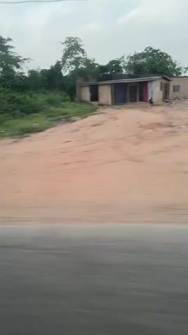 The video shows a view from a moving vehicle, looking out at a landscape of trees and scrub on a sandy embankment, adjacent to an asphalt road. The landscape is mostly green with some exposed soil. An abandoned or dilapidated building is visible in the background. The sky is overcast. The movement of the vehicle causes motion blur. Music is audible.