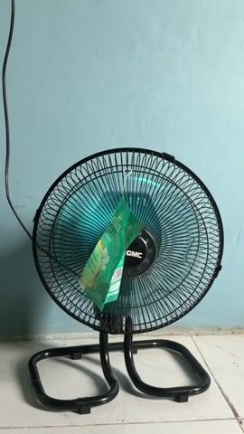 A black oscillating fan with a blue fan blade is positioned on a tiled floor in front of a light blue wall. A green sachet, possibly a scented sachet, is hanging from the front grill of the fan. The fan is plugged in, with a black cord visible extending upwards and to the left. The sachet has text and an image of flowers on it. The fan appears to be stationary in this frame.