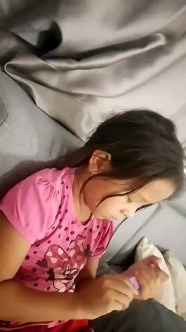 The video shows a young girl with dark hair wearing a pink t-shirt with polka dots and a Minnie Mouse design. She is lying down on a gray couch and holding a small purple object in her hands. Her gaze is directed downwards at the object. The background consists of the couch and some blurred fabric. The lighting is dim.