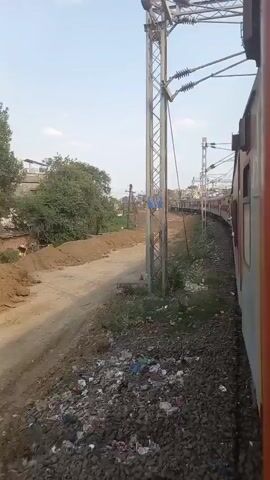 A train car, painted red and white, is moving alongside a second set of train tracks on the right of the frame. Next to the parallel tracks is a dirty, barren, dirt path alongside a patch of weeds and litter. Several metal and cement poles stand on the side of the tracks. The cement poles have blue plates with yellow numbers on them. Overhead is the wiring for the train. A city landscape is visible in the distance. The day is sunny.