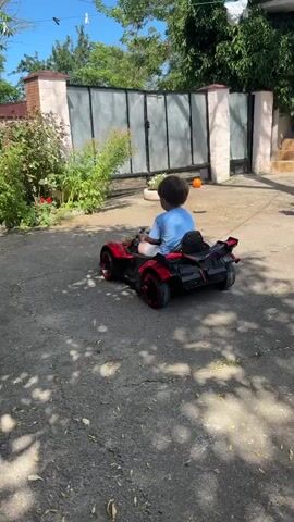 A child wearing a blue shirt is sitting in a black and red toy car. The child is facing away from the camera and appears to be driving the car forward. The car is on a paved surface, possibly a driveway or courtyard. In the background, there is a fence with large metal gates, green foliage, and part of a building. The sky is visible and appears to be blue and clear, suggesting it is daytime. There is a shadow of trees or other objects falling on the ground in front of the car. An orange ball is visible on the ground near the fence. The car has red accents on the wheels and body. The child's hands are on the steering wheel of the toy car. The overall scene depicts a child playing outdoors with a ride-on toy car.