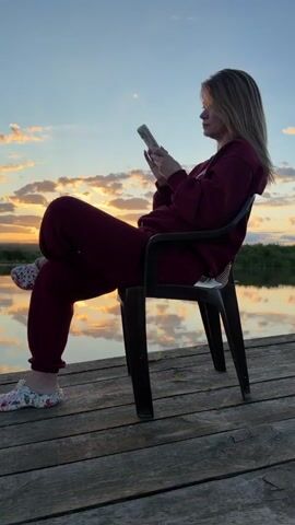 A young woman with blonde hair sits on a dark plastic chair on a wooden pier, looking at her phone. She is wearing a burgundy tracksuit and floral patterned slip-on shoes. The sun is setting over a calm body of water, casting reflections of the sky and clouds on its surface. The sky is filled with dramatic, orange-tinged clouds.
