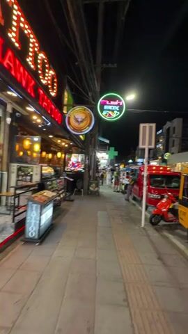 The video shows a nighttime street scene. In the foreground, a person wearing a patterned shirt walks away from the camera. To the left, a sidewalk features shops with illuminated signs and displays of food. To the right, several vehicles are parked, including a yellow tuk-tuk and a red truck. The street is lined with buildings and has streetlights. People are visible walking along the sidewalk in the background. Neon signs and lights illuminate the area, indicating a commercial district.
