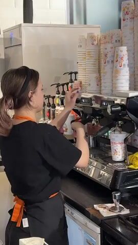 A young woman wearing a black t-shirt and an orange apron is working behind a coffee machine in a cafe. She has her hair pulled back in a ponytail and wears a black headband. In the video, she is seen placing two paper cups onto the coffee machine, then she appears to be making coffee. Stacks of paper cups are visible on a shelf above the coffee machine. A yellow cloth is draped over the coffee machine. There are various syrup bottles behind the coffee machine. The counter has a tamper and a portafilter on it.