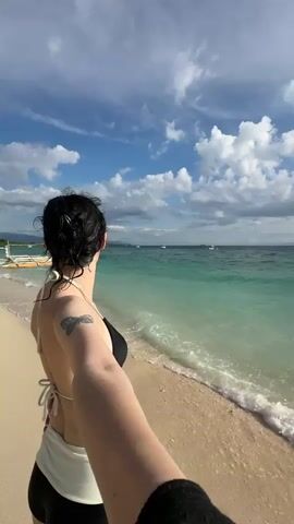 A woman with dark hair pulled back, wearing a black bikini top and white and black bottoms, stands on a sandy beach facing the ocean. She is holding a camera in her right hand, filming herself in a selfie-style shot as she looks out at the turquoise water. Gentle waves lap the shore, and the sky is blue with scattered white clouds. In the distance, several small boats are visible on the water. The beach appears to be lined with palm trees and some structures on the left side of the frame. The video pans slightly, offering a wider view of the beach and the ocean.