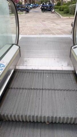 The video opens from the perspective of someone standing on an escalator, looking down. The escalator's metal steps are prominent in the foreground, suggesting an entrance or exit. As the view expands, it reveals a lively outdoor scene, likely a promenade or town square in Kotor, Montenegro.

Palm trees are a dominant feature, their fronds swaying gently, indicating a warm or temperate climate. In the background, rugged, tree-covered mountains rise majestically, a characteristic feature of the Kotor region. Scattered among the trees and along the paved walkway are several parked scooters or small vehicles, suggesting transportation options available to visitors or residents.

People are visible walking and interacting, creating a sense of normalcy and activity. Some are seen in small groups, while others are alone. The lighting suggests it's either late afternoon or early evening, with soft daylight filtering through the palm trees. The sky appears partly cloudy.

The paved ground is laid with decorative stone tiles, adding to the aesthetic appeal of the area. Benches are also visible, offering places for people to rest and enjoy the surroundings. The overall atmosphere appears relaxed and pleasant, characteristic of a tourist destination or a well-maintained public space. No visible text is present in the video.