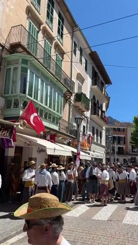 The video shows a festival in a town square with a large stone cathedral in the background. Many people in historical costumes fill the area, some carrying flags. There are buildings with balconies on either side of the square. The ground is paved with red bricks in a herringbone pattern.  Tram lines run through the square, and a pole is decorated with a red ribbon labeled "BATLLA." The sky is clear blue. There are some cannon shots.