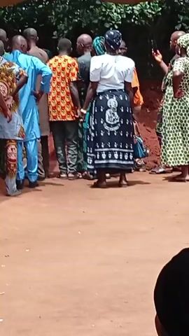 The video shows a gathering of people, apparently outdoors on a patch of reddish soil. The group is diverse in age and attire. Many women wear colorful wraps or skirts. Some individuals wear traditional clothing, while others opt for modern apparel. Interactions include people talking and generally being in close proximity. A few of the women are wearing wraps with an image of the Virgin Mary on them. The video transitions to a view of people near the base of a house. The focus shifts between wider group shots and closer views of smaller clusters of individuals. Overall, the video captures a scene of community and social interaction.