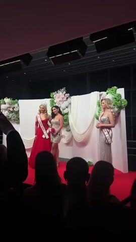Three women stand on a red carpeted stage in front of a crowd. The woman on the left wears a red gown and a crown, with a sash across her chest. In the center stands a woman in a short, sparkly dress and heels, holding a rose. To the right, a woman in a long, silvery gown holds a bouquet of pink roses. Each woman wears a sash. Behind them is a backdrop of white panels decorated with white drapery and arrangements of pink and white flowers. The audience sits facing the stage, their silhouettes visible in the foreground. Above, dark lighting fixtures hang from the ceiling. Large windows are visible in the background.