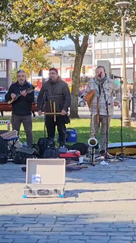 A group of four men are performing as a street band in a public area. One man is playing an eight-stringed mandolin-like instrument, another is singing and playing a double recorder, a third is playing panpipes and another one is playing guitar. In front of them is a closed silver-colored briefcase, likely for tips. A saxophone is on the ground between two of the musicians. They are standing on a stone-paved area with a tree behind them. In the background are buildings and a street with passing cars. The scene is bright and sunny.
