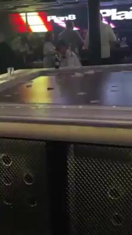 The camera focuses on a bar with a rectangular, lit-up top, flashing through red, blue, and purple.  Around the bar, numerous individuals stand in the background, indistinct due to the lighting and distance. The base of the bar is dark, textured, and punctuated with small, circular indentations. The lighting is predominantly dark with flashing, colorful effects.