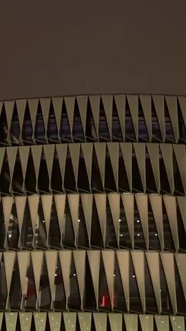 This is a vertical, close-up, exterior shot of a building at night. The building has a modern architectural design with repeating triangular elements forming horizontal bands. The triangular elements are angled, with some angled inwards and some angled outwards, creating a layered effect. The lighting suggests it is nighttime, with a dark sky and interior lights visible through the gaps in the triangular facade. The overall tone is somewhat muted and atmospheric due to the low light conditions and the cool color palette of the sky and building materials. There are no people or distinct objects visible in the frame, only the architectural structure itself.