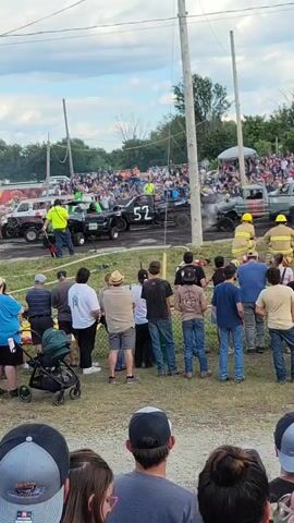 The video shows a demolition derby event outdoors. In the foreground, a crowd of spectators stands watching. In the middle ground, two wrecked pickup trucks are positioned on a dirt track. One truck is black with the number 52 visible on its side, and the other is greenish-gray. There is smoke emanating from the greenish-gray truck. Several individuals in yellow and red safety gear, likely safety personnel or firefighters, are near the greenish-gray truck. On the left side of the track, a man in a bright yellow vest is standing near another wrecked vehicle. In the background, a large crowd of spectators is gathered behind barriers, and tents and buildings are visible. The sky is partly cloudy. The video appears to be filmed from a slight upward angle, looking towards the derby track.