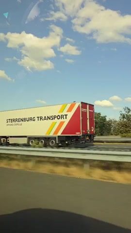 A white semi-trailer truck with "STERRENBURG TRANSPORT" and "SPRANG-CAPELLE" printed on its side travels on a highway under a blue sky with white clouds. The truck has a red and colorful striped design on its rear. The shot is taken from a moving vehicle, capturing the truck from the side. The environment includes a highway barrier and trees in the background.