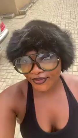 A woman is facing the camera in an outdoor setting. She is wearing a black tank top, a large pair of tinted aviator sunglasses, and a large furry black hat. The background consists of a brick-paved street and houses. The woman is smiling slightly. The camera is held at a close distance and appears somewhat shaky.