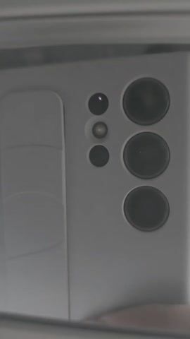 The video shows the back of a smartphone with multiple cameras. The smartphone is white or light gray and is being held in someone's hand, reflected in a mirror. There are three large camera lenses and three smaller ones positioned above them. The phone is stationary. There is a light sound in the background.