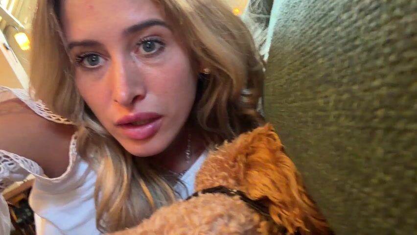 A woman with blonde hair and blue eyes is holding a small, curly-haired, reddish-brown dog. She asks the dog to wake up. The dog is lying on a green couch. The camera angle shifts to show the back of the dog's head, then its face as it looks around. A potted plant, a wooden table, and chairs are visible in the background.