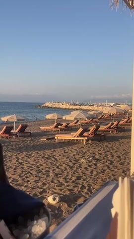 The video opens with a close-up view of two dark glass bottles nestled in an ice bucket on a sandy beach. The bottles are positioned in the foreground, with one bottle slightly obscuring the other. The bucket contains ice and some white paper elements. The background reveals a tranquil beach scene under a clear blue sky. Numerous empty lounge chairs and umbrellas are arranged on the sand, facing the calm ocean. A breakwater or pier extends into the sea in the distance, and a few people can be seen swimming or wading in the water. The lighting suggests it is either early morning or late afternoon, with a gentle, warm glow. The overall atmosphere is one of relaxation and leisure.