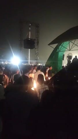 A nighttime gathering of people illuminated by bright lights and a speaker system suspended from a metal truss. The crowd is dense, with many individuals holding small, lit objects, possibly candles or lanterns, which cast a warm glow. A stage structure with a green awning is visible on the right side of the frame. The overall atmosphere appears to be festive or commemorative.