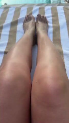 The video shows a first-person perspective of a person lying on a striped towel by a swimming pool. The person's legs and feet are visible, with their legs extended towards the pool. The water in the pool reflects trees and possibly buildings. The lighting suggests it is daytime, likely sunny, as the legs appear to be catching the light. The scene is static, with no noticeable movement beyond slight shifts in the camera's angle.