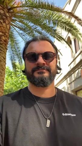 A man with dark hair, a beard, and sunglasses is shown from the chest up, standing outdoors. He is wearing a black sweatshirt with a pendant necklace. Behind him, a palm tree is visible on the left, and a building with arched windows and a "GUCCI" sign is on the right. The man is looking at the camera and appears to be talking. The lighting suggests it is daytime.