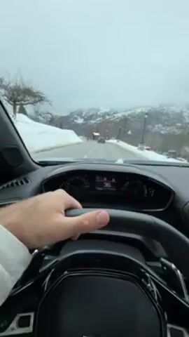 A first-person perspective inside a car driving on a snow-covered mountain road. The driver’s left hand is visible on the steering wheel. The dashboard shows a digital speedometer with a speed fluctuating between 36 and 31 km/h.  The car's infotainment system displays a navigation map. Snow is on the sides of the road, and mountains are in the background. There are other cars on the road. The weather is overcast and cloudy. A conversation is audible in the background. The camera pans slightly from side to side, showing more of the car's interior and the surrounding environment.