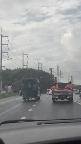 This video captures a view from inside a vehicle looking out at traffic on a wet road. In the foreground, the dashboard of the vehicle is visible. Ahead, a blue three-wheeled vehicle, similar to a tuk-tuk, is driving with several people in the back. To the right of the blue vehicle, a silver pickup truck is in motion. Several individuals are riding in the bed of the pickup truck, some wearing orange shirts. The pickup truck has a sticker on the back with Thai writing and a phone number. In the background, there are trees, utility poles with wires, and telecommunication towers. The sky is overcast with clouds. The video appears to be taken while the vehicle is moving through traffic.