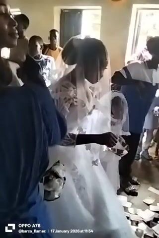 The video depicts a bride in a white wedding dress, accompanied by a child also in white, walking through a room where money is being thrown in the air and scattered on the floor. Several people are in the background, some standing, watching the bride. A person in a white headwrap can be seen helping adjust the bride's dress, picking up money from the floor. The setting seems to be indoors, likely a celebration venue or religious building. The video is taken with a smartphone, as indicated by the "OPPO A78" watermark.