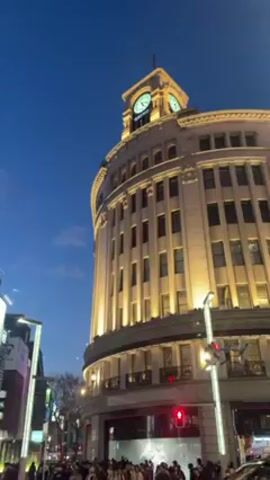 The video shows a bustling street scene in Japan at dusk. The iconic Wako building with its clock tower is prominently featured. Numerous pedestrians are crossing the street at a crosswalk. Cars, including an Uber and a taxi, navigate through the intersection. The sky is a deep blue, transitioning from daylight to evening. Buildings with illuminated advertisements rise in the background. The scene is captured with a slightly upward angle, showcasing the cityscape and the movement of people and vehicles.