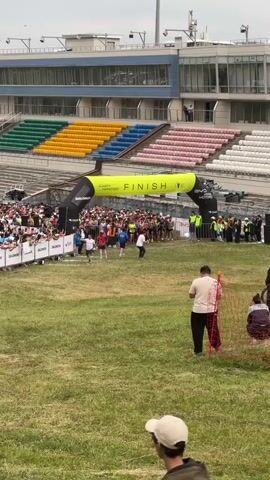 The video begins with a view of a starting or finishing line for a race. A large inflatable archway with "FINISH" written on it is visible, with a crowd of runners behind it. Spectators are seated in bleachers in the background. A person on a motorcycle is seen riding across the frame from left to right, passing in front of the runners. The grass in the foreground is green and appears to be a racecourse. The sky is overcast. The camera is positioned at ground level, looking up towards the archway and the crowd. The image is slightly blurry and appears to be a low-resolution video.
