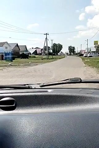 The video shows a view from inside a vehicle, looking out through the windshield at a paved road in a rural area. The road is lined with houses and trees. There are power lines running along the side of the road. Several cars are parked along the side of the road. A speed limit sign is visible on the right side of the road. The sky is partly cloudy and the sun is shining. The video appears to be taken while the vehicle is in motion.