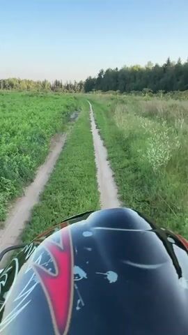 The video shows a first-person perspective of someone riding an ATV on a dirt path through a grassy field. The sky is clear and blue. There are trees in the distance. The ATV has a black helmet with red and white designs on it. The path is narrow and has some muddy patches. The ATV is moving forward along the path.
