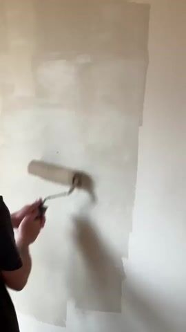 A man with a beard and tattoos on his arm is painting a wall with a paint roller. The wall is half painted with a light color, and the other half is a darker, unpainted color. The man is wearing a dark shirt. The room is well-lit, and the wall appears to be indoors.