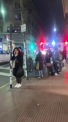 The video shows a street scene at night. A fire truck with flashing red lights is visible in the background. Several people are standing on the sidewalk, some of whom appear to be looking at their phones. A woman in the foreground is speaking on her phone. The scene appears to be a city street with buildings and traffic lights. The overall mood is calm.
