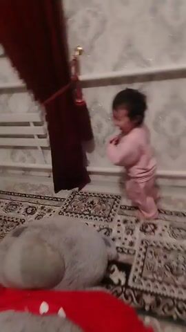 A young child, dressed in a pink jumpsuit, is walking on a rug in a room. The child is holding a small red object in their hand. The room has patterned wallpaper, a window, a radiator, and a small white cabinet. The child appears to be happily running towards the camera and then to the side. The child interacts with the cabinet, leaning on it for support and smiling. The child makes a noise.